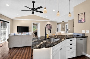 A modern kitchen with a central island, a ceiling fan, pendant lights, and a large flat-screen TV.