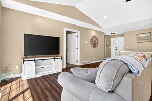 A living room with a flat-screen TV, a gray sofa, wooden flooring, and a door leading to another room. - Fall Getaway Base Camp: Wine Nights & Easy Access to Silver Lake State Park (Shelby)