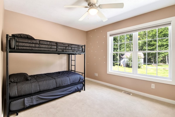 A room with a bunk bed, a ceiling fan, a large window with a view of greenery, and a carpeted floor.
