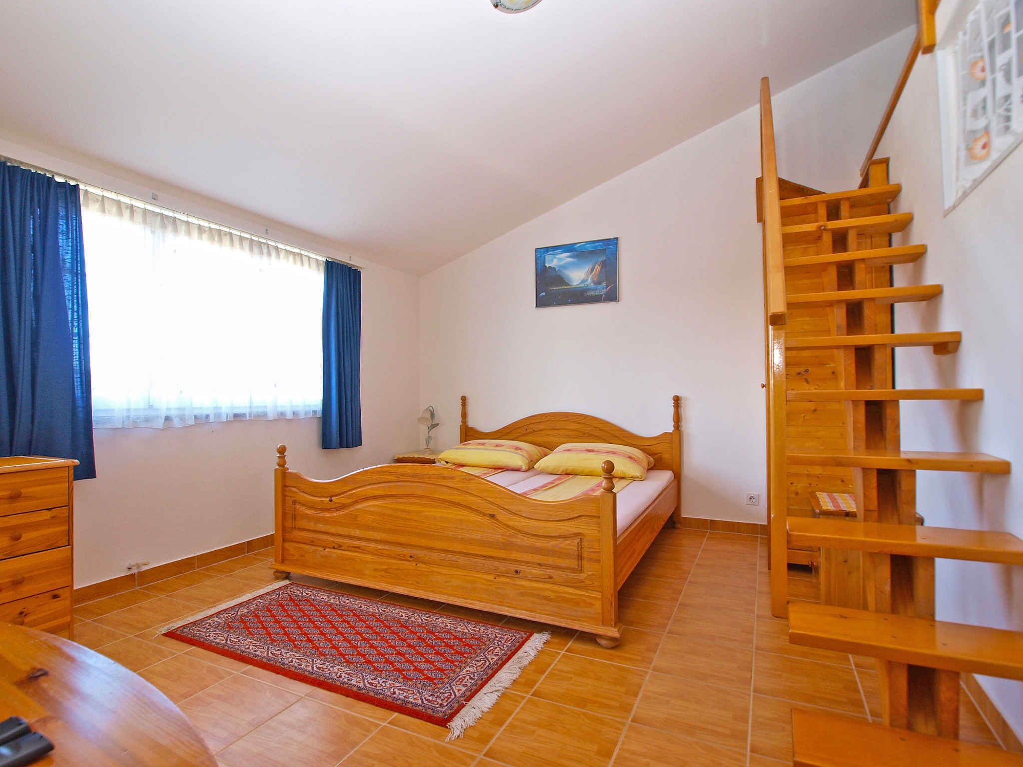 A bedroom with a wooden bed, a red patterned rug, a wooden dresser, a window with blue curtains, and a wooden staircase.