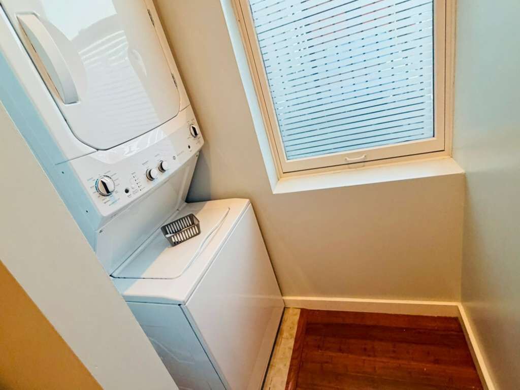 Un área de lavandería con lavadora y secadora, una ventana con persianas y piso de madera.