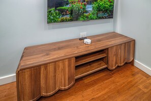 A wooden console table with a built-in TV screen displaying a garden scene, a power outlet, and a remote control. - Central OTR, Lux King Bed, 2 Smart TVs, Sleeps 3ppl, Fast Wifi, 1st Floor (Cincinnati)