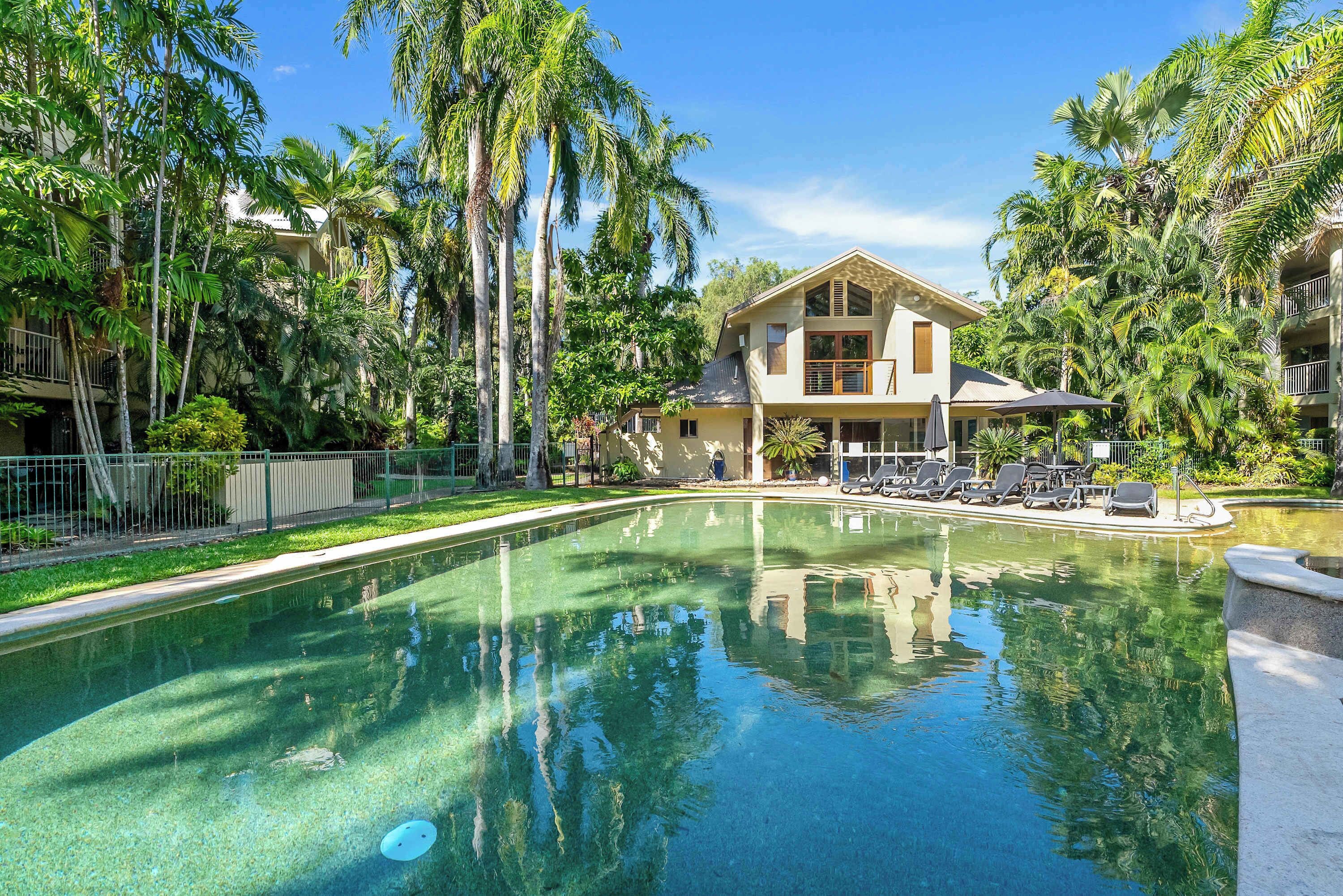 A pool with clear water, surrounded by lush greenery and a two-story building with balconies.