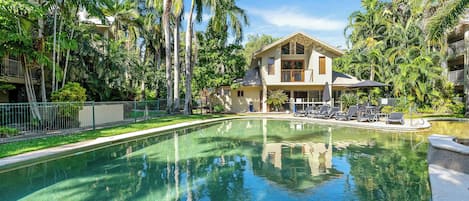 A pool with clear water, surrounded by lush greenery and a two-story building with balconies.