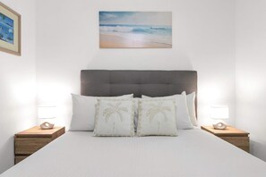 Ein Schlafzimmer mit einem Bett, zwei Nachttischen, zwei Lampen, einem Kopfteil und einem Bild von einer Strandansicht.