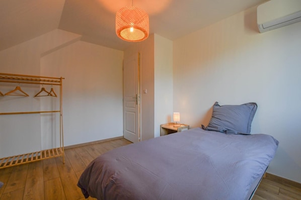 A bedroom with a bed, a hanging light fixture, a bedside table with a lamp, and a wooden floor.