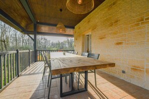 Een terras buiten met een houten tafel en stoelen, een stenen muur en uitzicht op bomen.