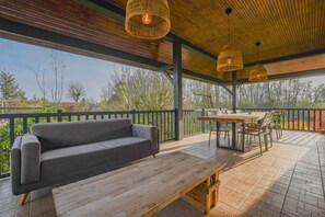 Een terras buiten met een houten tafel en stoelen, een grijze bank en hangende rieten lampen.