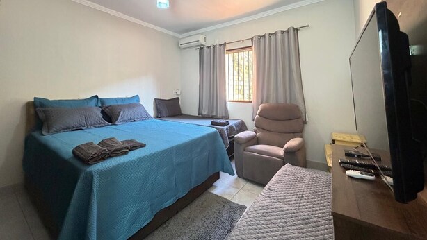 Un dormitorio con cama, una silla, un escritorio con televisor y una ventana con cortinas.