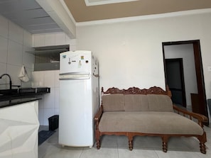 A compact living space with a kitchenette, a white refrigerator, a wooden sofa, and a door leading to another room.