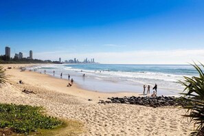 Uma praia com pessoas, um horizonte urbano ao fundo e céu claro.