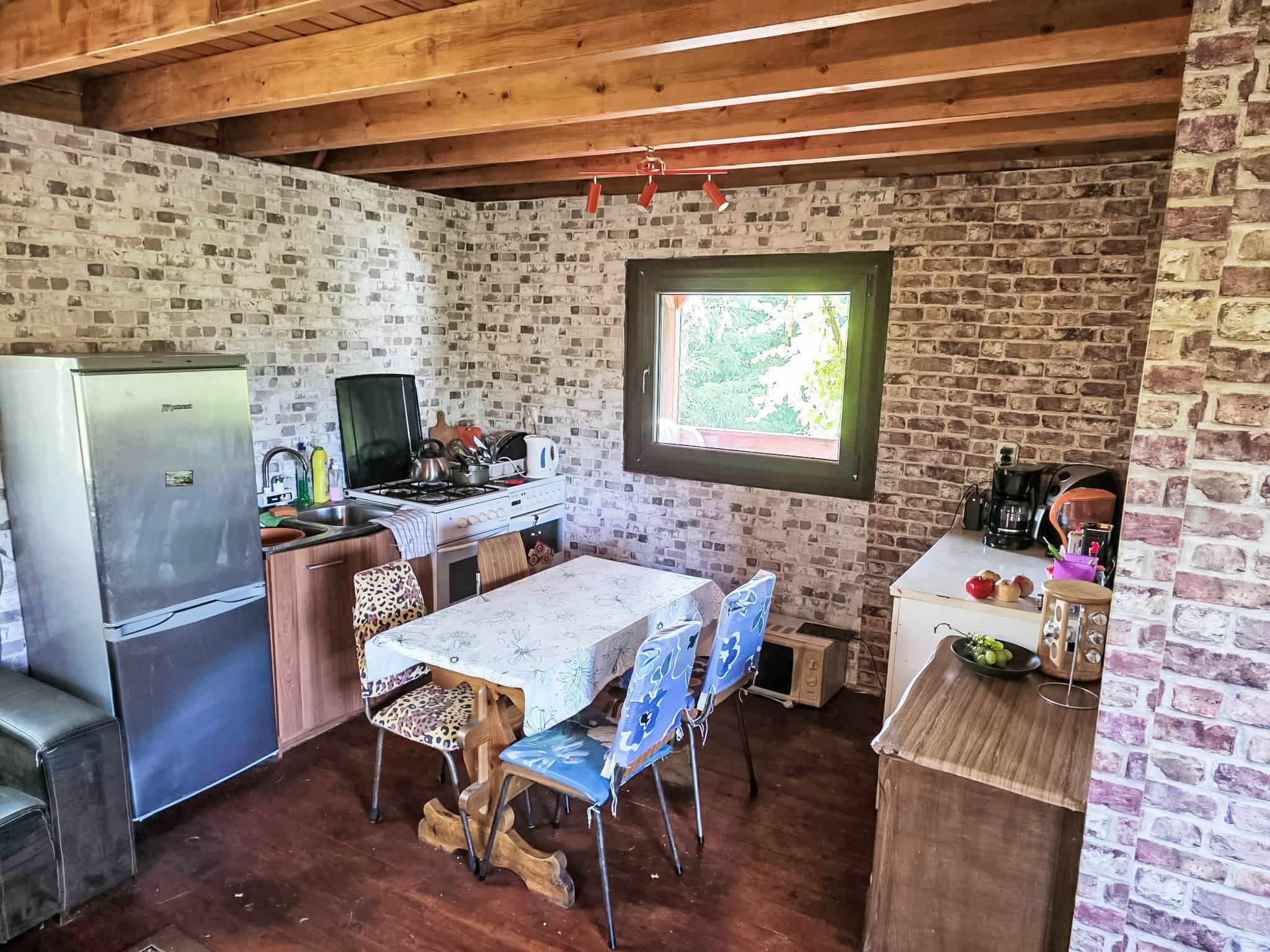 Een landelijke keuken met een bakstenen muur, houten balkenplafond, een eettafel met stoelen, een koelkast, een fornuis en een raam.