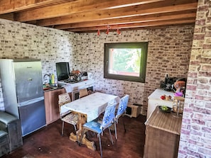 Una cocina rústica con pared de ladrillo, vigas de madera en el techo, una mesa de comedor con sillas, un refrigerador, una estufa y una ventana.