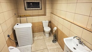 Casa de banho compacta com sanita, máquina de lavar roupa, lavatório e janela sobre a sanita.