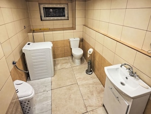 Een kleine badkamer met toilet, een wasmachine, een wastafel en een raam boven het toilet.