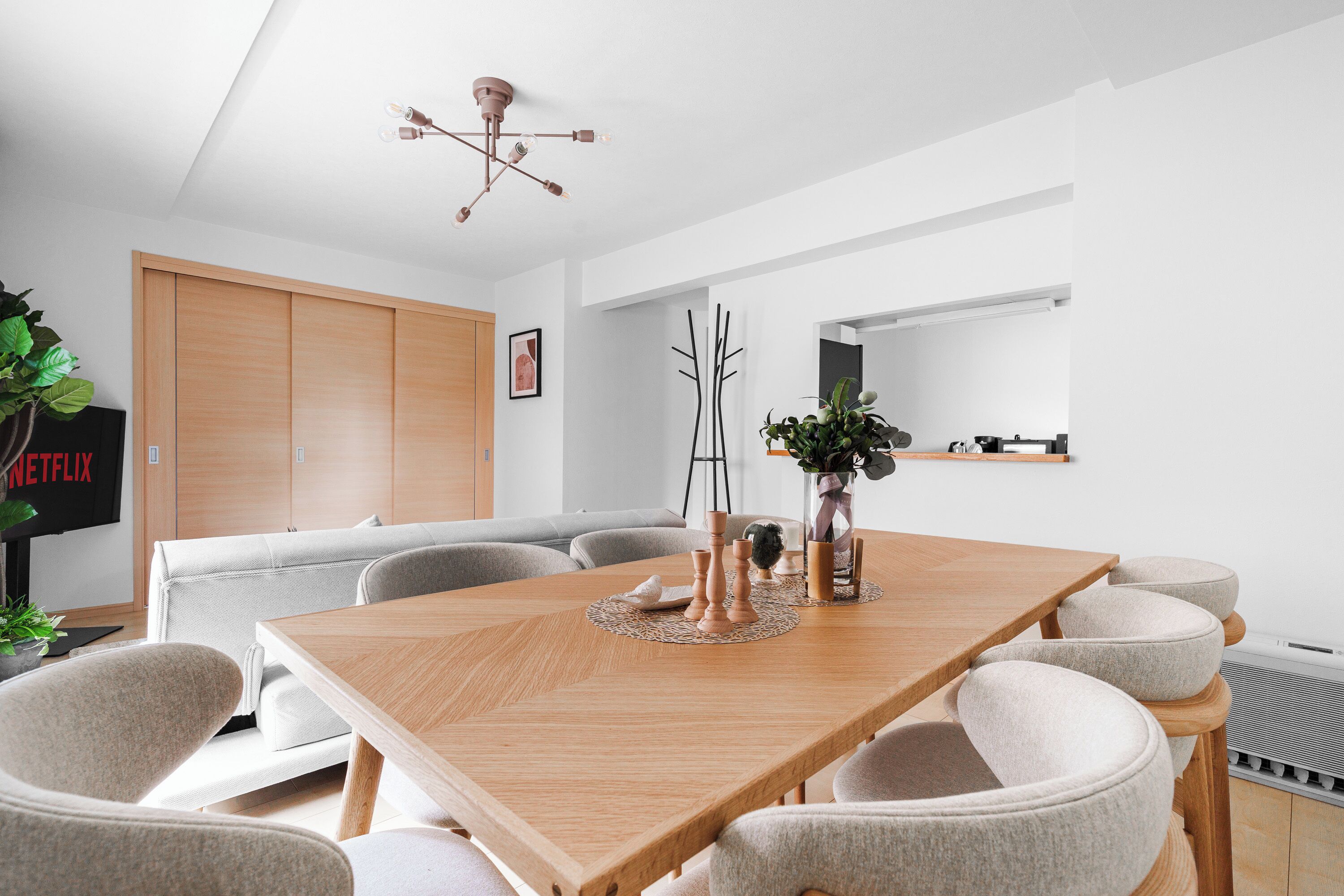 A modern dining area with a wooden table, grey upholstered chairs, a ceiling light fixture, and a plant on the table.