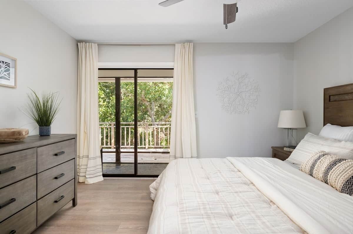 A bedroom with a bed, a dresser, a nightstand, a lamp, and a view of a balcony with a railing.