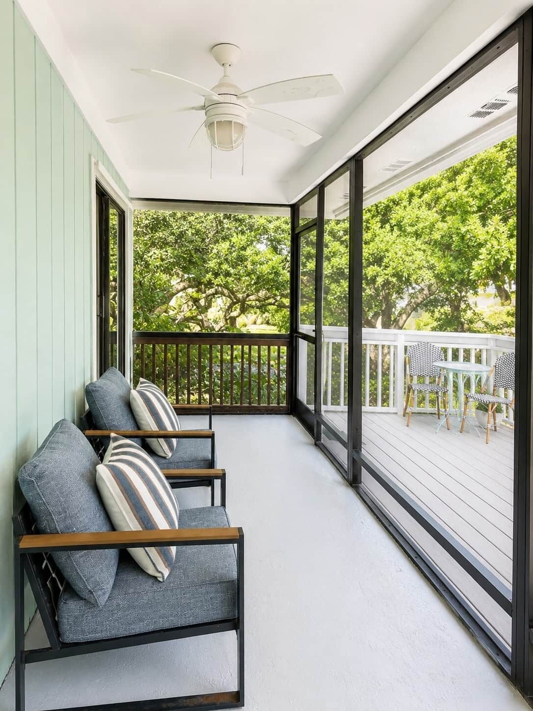 A covered porch with a ceiling fan, a seating area with cushioned chairs and pillows, and a view of a tree-lined yard.