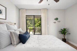 A bedroom with a bed, curtains, a ceiling fan, a window with a view, and a potted plant. - Palmetto Place | Beach, Pool & Walkable Dining (Vero Beach)