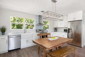 A modern kitchen with white cabinetry, a stainless steel refrigerator, a wooden dining table, and a built-in dishwasher. - Palmetto Place | Beach, Pool & Walkable Dining (Vero Beach)