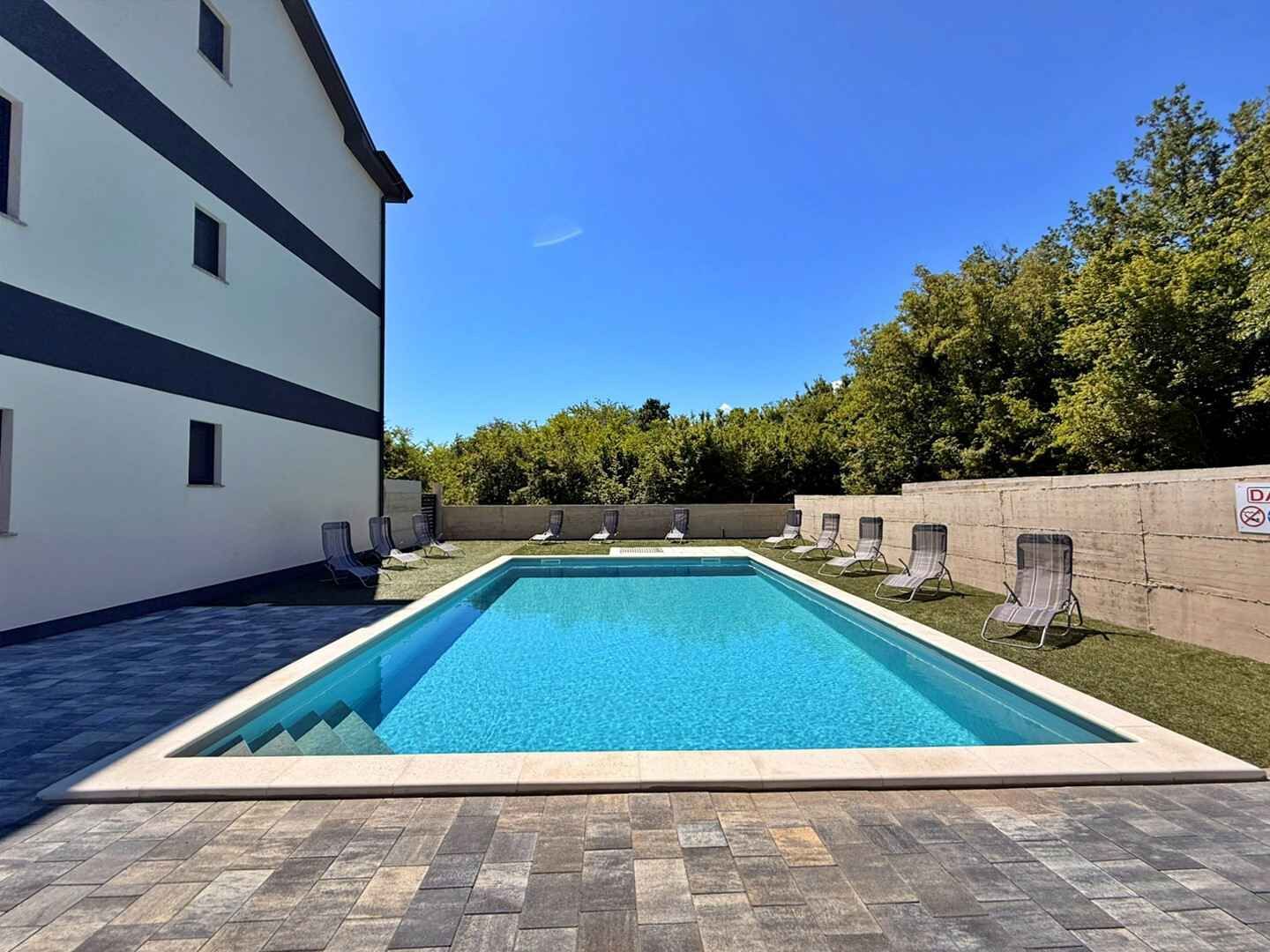 A rectangular swimming pool with a tiled deck, surrounded by lounge chairs and a building with multiple windows.