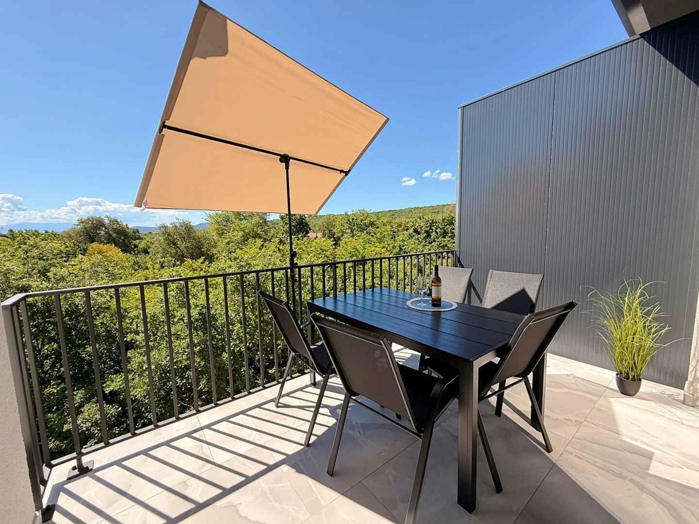 Uma varanda com mesa e cadeiras, um guarda-sol de mesa e um vaso com planta.