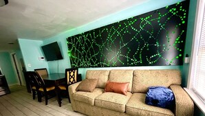 A living room with a beige sofa, a dining table with chairs, and a wall-mounted TV.