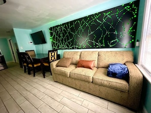 Ein Wohnzimmer mit einem beigen Sofa, einem Esstisch mit Stühlen und einem an der Wand befestigten Fernseher.