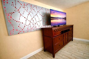 A room with a flat-screen TV on a wooden cabinet, a large abstract painting on the wall, and a herringbone-patterned floor. - Glow Hard or Go Home - 12 mins to Downtown (Jacksonville)
