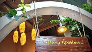 Un letrero de madera con el texto 'Tiffany Apartment' colgado de una estructura blanca curva, con luces colgantes y follaje.