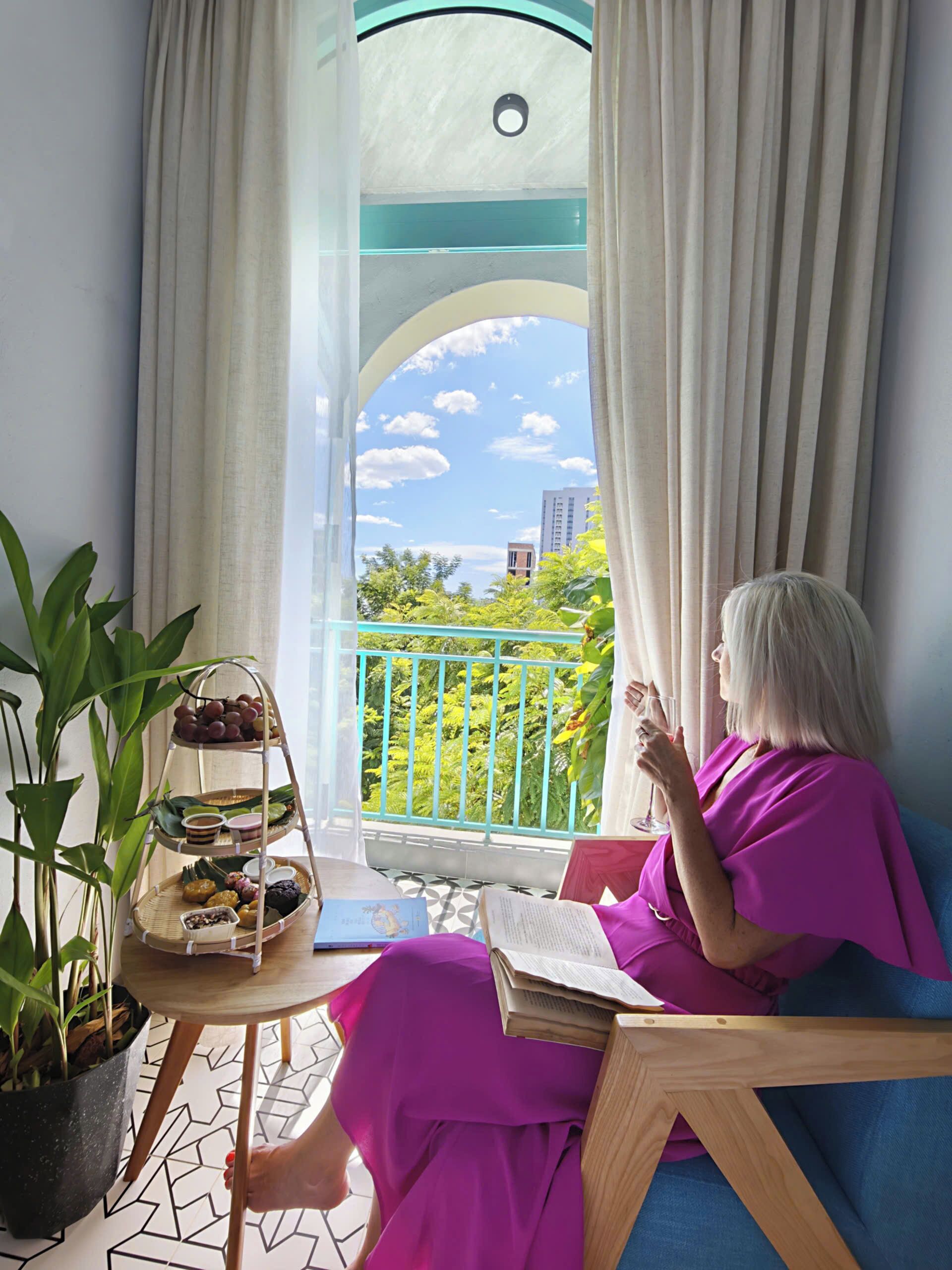 Una mujer sentada en una silla de balcón, leyendo un libro, con una bandeja escalonada de aperitivos y una planta en maceta a su lado.