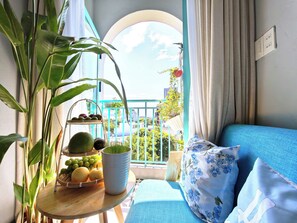 Un balcón con vista a edificios, una mesa con frutas y una planta en maceta, y un sofá azul con cojines estampados.