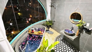 Un baño moderno con una bañera grande, ducha, felpudo de baño colgado y vista de la ciudad por la noche.