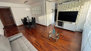 Een moderne woonkamer met een houten vloer, een grijze bank, een eettafel met stoelen, een flatscreen televisie en een decoratief wandpaneel.