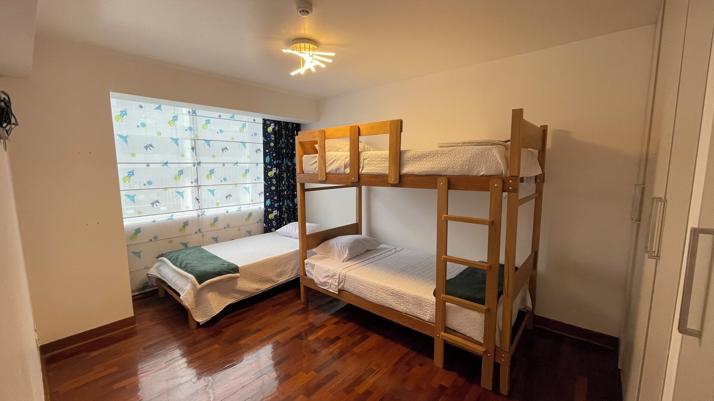 A room with bunk beds, a window with patterned curtains, and a ceiling light fixture.