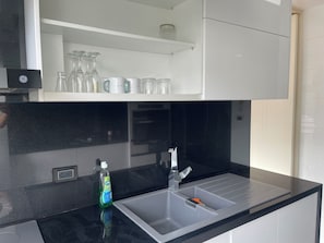 Une cuisine avec un plan de travail noir, un évier en acier inoxydable et une étagère au-dessus, où sont rangés des verres et un bocal.