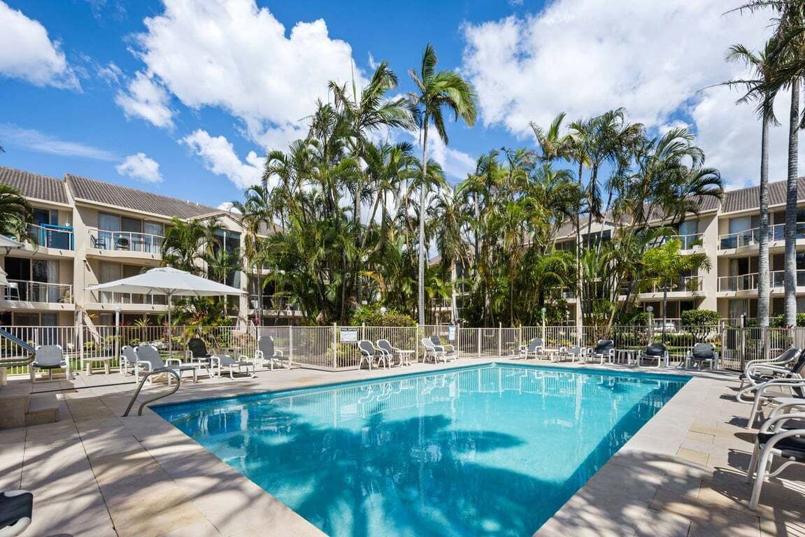 Una piscina con lettini, circondata da palme e dall'edificio di un hotel.