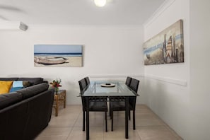 A dining area with a glass table and chairs, a sofa with cushions, and two wall-mounted paintings. - Miami Beachside - 32 (Gold Coast)