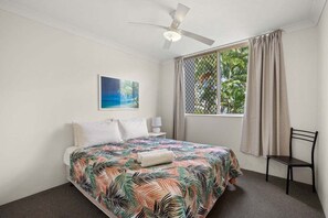 Ein Schlafzimmer mit einem Bett, einem Deckenventilator, einem Fenster mit Vorhängen, einem Sessel und einem an der Wand hängenden Bild.