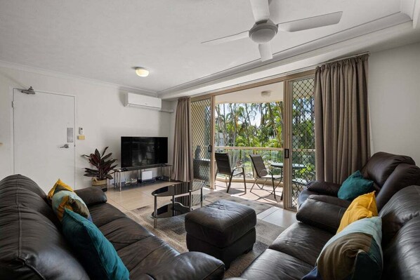 Ein modernes Wohnzimmer mit Ledersofa, Flachbildfernseher, Deckenventilator und einer Glastür, die auf einen Balkon mit Gartenmöbeln führt.