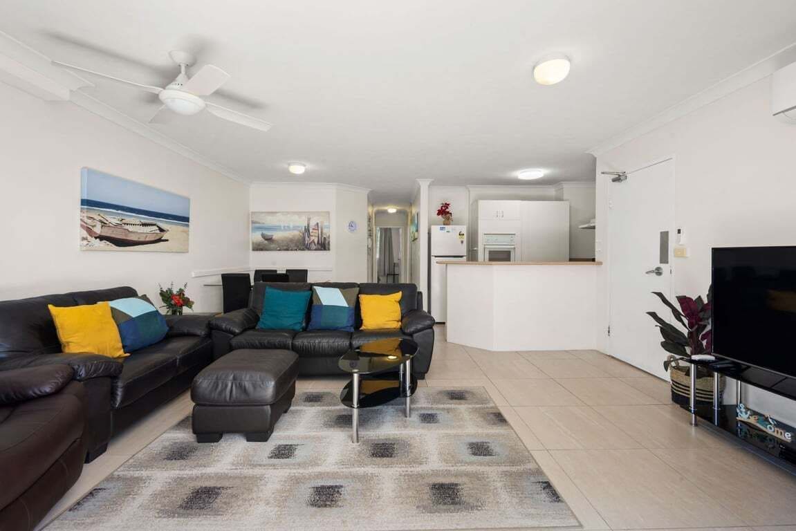 A modern living room with a sofa, ottoman, and coffee table. There is a television, a ceiling fan, and artwork on the walls.