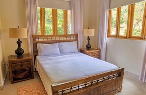 A bedroom with a wooden bed, two bedside tables with lamps, a nightstand with a drawer, and a window with curtains. - Vrbo Property (Jarabacoa)