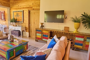 A cozy living room with a wooden ceiling, a large flat-screen TV mounted on the wall, a colorful cabinet with drawers, a sofa with cushions, and a small table with decorative items.