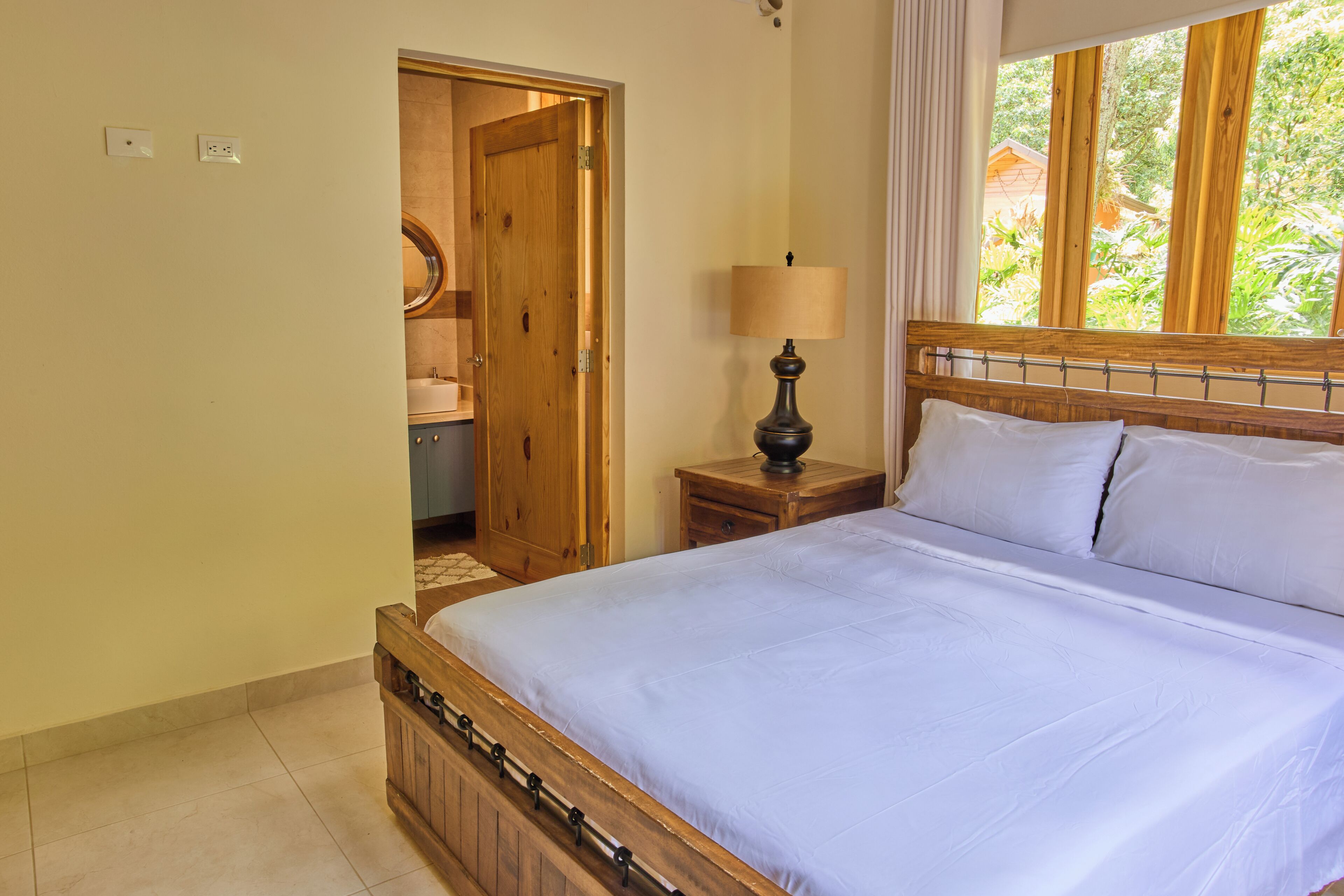 A bedroom with a wooden bed, a bedside table with a lamp, a window with curtains, and a door leading to a bathroom.