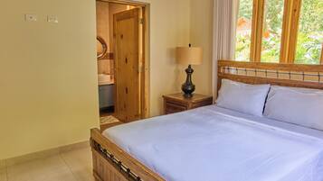 A bedroom with a wooden bed, a bedside table with a lamp, a window with curtains, and a door leading to a bathroom.