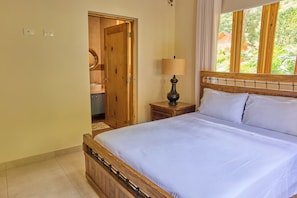 A bedroom with a wooden bed, a bedside table with a lamp, a window with curtains, and a door leading to a bathroom.