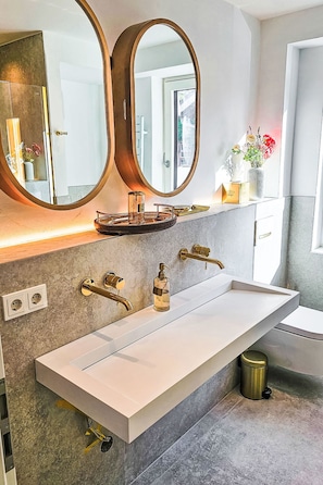 Ein modernes Badezimmer mit einem weißen Waschbecken, zwei goldenen Wasserhähnen und einem großen Spiegel darüber.