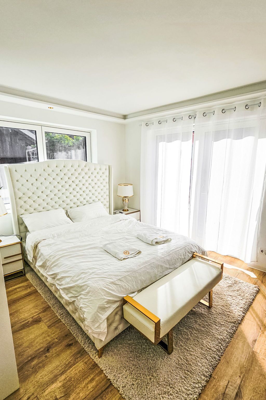 Ein Schlafzimmer mit einem Bett, Nachttischen, einer Bank, einem Fenster mit Vorhängen und Blick ins Freie.