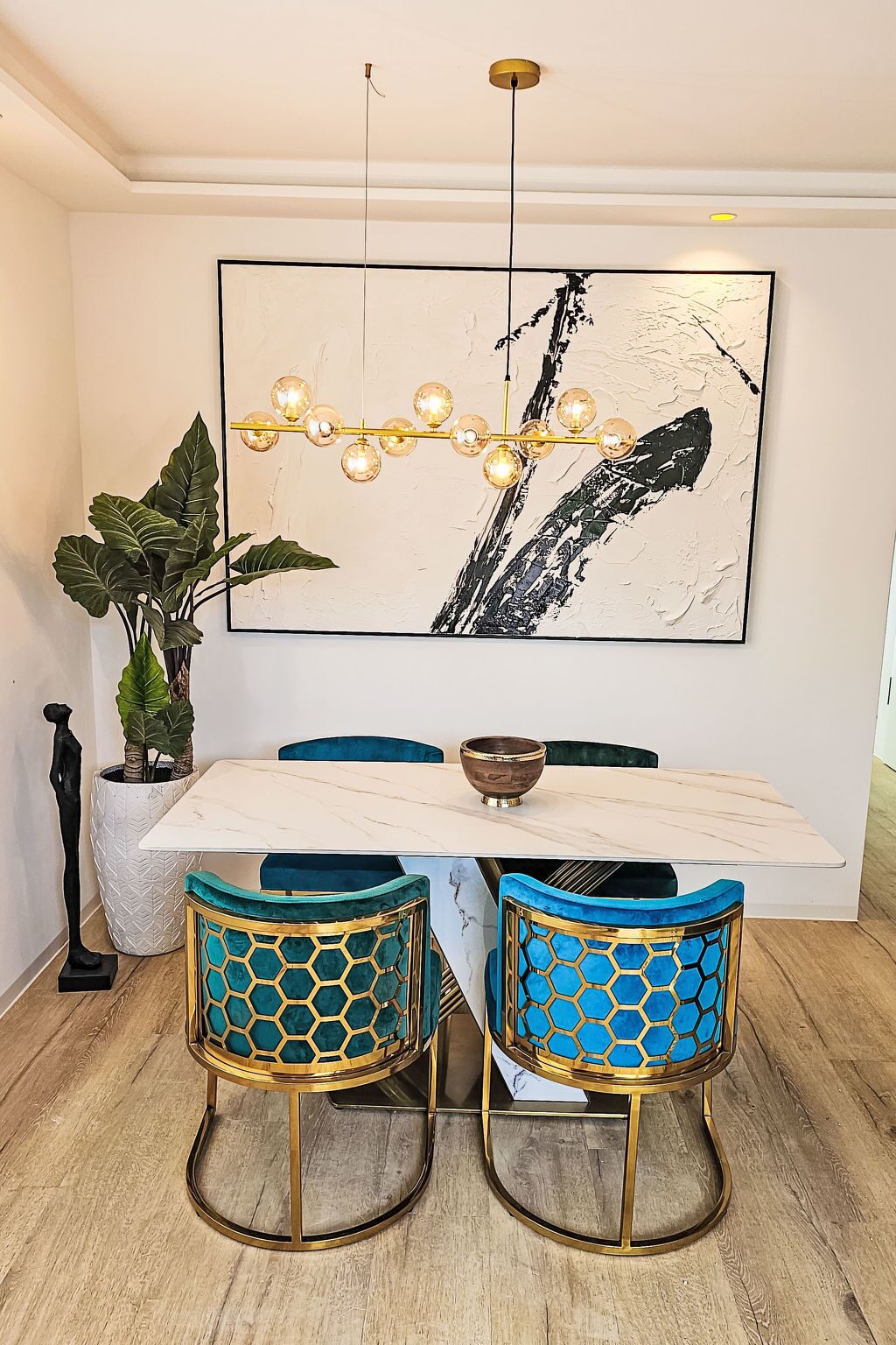 A dining area with a white marble table, two blue chairs with gold and teal accents, a large abstract painting, and a hanging light fixture.