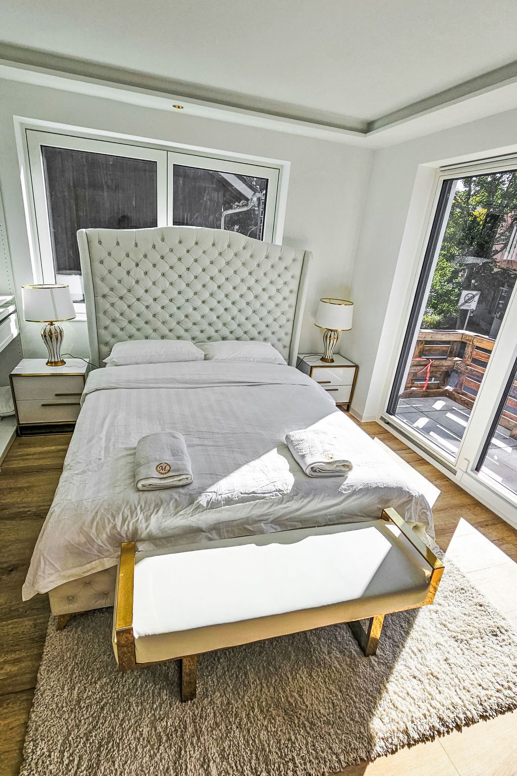 A bedroom with a large bed, two bedside tables with lamps, a bench at the foot of the bed, and a view of the outside through a large window.